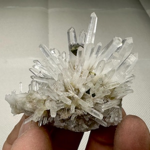 May include: A cluster of clear quartz crystals, with numerous pointed formations. The crystals are grouped together, with larger points in the centre. The base is a rough, earthy colour, contrasting with the transparent crystals. The quartz is a natural mineral.