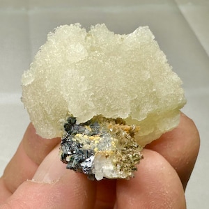 Może przedstawiać: Jasnożółty okaz minerału o krystalicznej strukturze. Górna część to większa, teksturowana formacja, a podstawa to mieszanka ciemnych i jasnych minerałów. Okaz trzymany jest w dłoni.