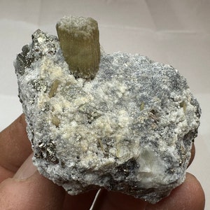 Puede incluir: Una roca gris rugosa con un gran cristal marrón claro en la parte superior. La roca está cubierta de cristales blancos más pequeños y tiene un brillo metálico.