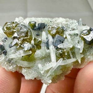 Może przedstawiać: Zbliżenie okazu mineralnego z skupiskami oliwkowozielonych kryształów, przezroczystymi końcówkami kwarcu i ciemnymi, metalicznymi akcentami. Minerał osadzony jest na jasnej, teksturowanej podstawie.