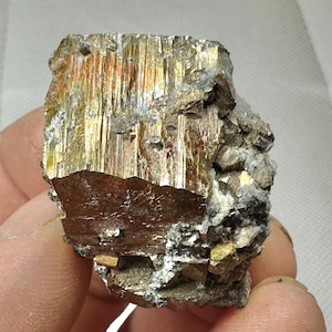 Op de afbeelding: Een close-up van een pyrietmonster, ook bekend als 'fool's gold'. Het mineraal vertoont een metaalglans met een mix van goud, brons en zilver tinten. De kristalstructuur is zichtbaar, met een gelaagde uitstraling. Het monster wordt in een hand gehouden.