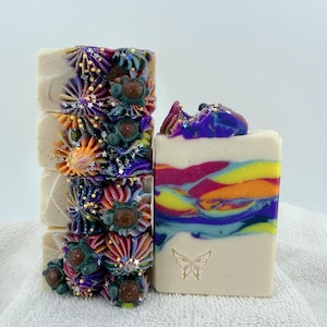 May include: A stack of four rectangular bars of soap topped with purple, orange, and blue piped frosting, small brown spheres, and tiny star-shaped glitter. A single bar stands beside them, featuring a colorful swirl and a butterfly stamp.