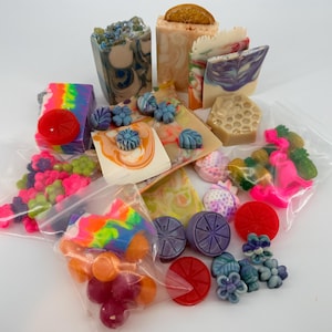 May include: A collection of assorted handmade soaps and wax melts in various shapes, colors, and sizes. The set includes rectangular bars with swirls, honeycomb patterns, floral accents, fruit-shaped pieces, and small items packaged in clear plastic bags.