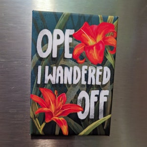 Puede incluir: Un imán verde, rojo y blanco con el texto "Oops I wandered off" y dos flores de lirio rojas.