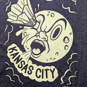 May include: A black and white illustration of a cartoon moon with a surprised expression. The moon is being hit by a badminton shuttlecock. The text "KANSAS CITY" is written in a cloud below the moon.