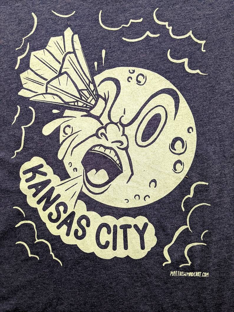 May include: A black and white illustration of a cartoon moon with a surprised expression. The moon is being hit by a badminton shuttlecock. The text "KANSAS CITY" is written in a cloud below the moon.
