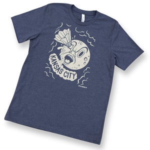 May include: A navy blue t-shirt with a white graphic of a cartoon moon with a surprised expression blowing a cloud of smoke that says "KANSAS CITY". The moon is holding a badminton racket and shuttlecock.