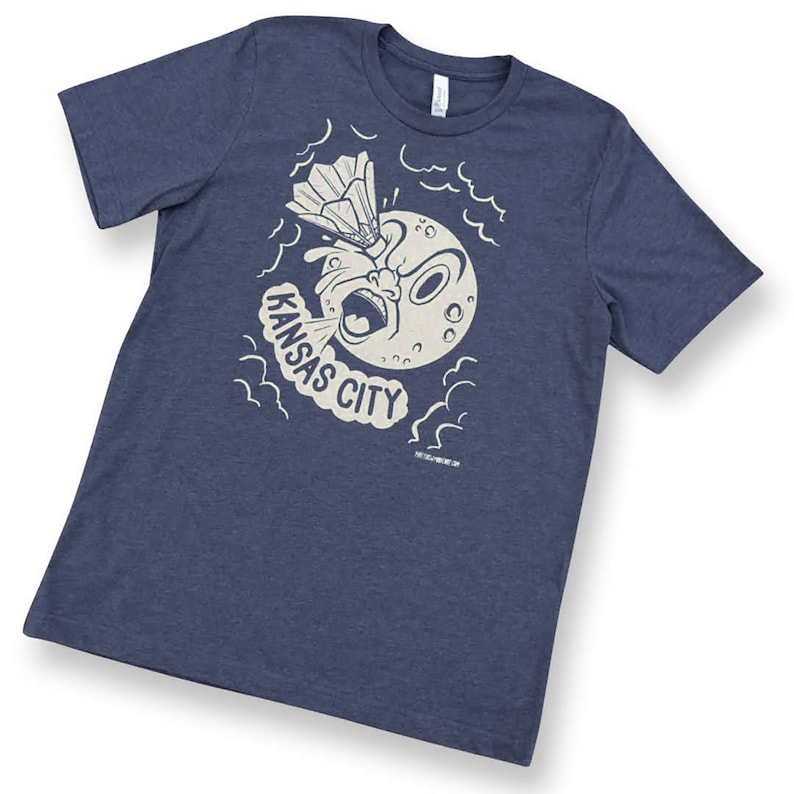 May include: A navy blue t-shirt with a white graphic of a cartoon moon with a surprised expression blowing a cloud of smoke that says "KANSAS CITY". The moon is holding a badminton racket and shuttlecock.