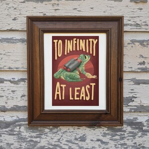 May include: A framed print with a green frog wearing a rocket pack. The text on the print reads "To Infinity At Least".