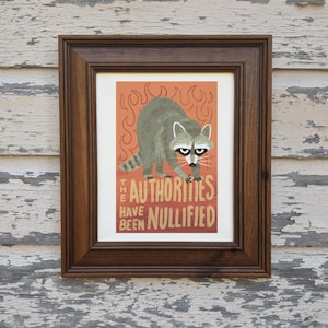 May include: A framed print with a raccoon illustration and the text "The Authorities Have Been Nullified". The raccoon is standing on its hind legs with its front paws raised. The background is a bright orange with flames.