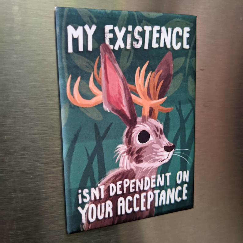 May include: A green and brown illustrated magnet with a rabbit with antlers. The text on the magnet reads "My existence isn't dependent on your acceptance."