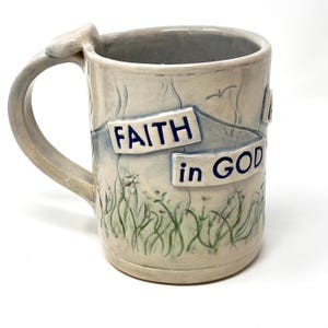 May include: A ceramic mug with a light grey glaze featuring a hand-sculpted handle and raised rectangular plaques with the dark blue text FAITH in GOD. The surface is decorated with painted green grass, faint mountain outlines, and a small bird.