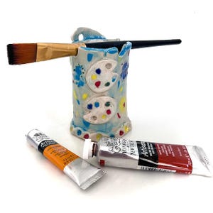 May include: A ceramic paintbrush holder with a light blue base, decorated with colourful paint palettes and floral designs. Two paintbrushes rest in the holder, and two tubes of paint are in front of it. One tube is orange, and the other is red.