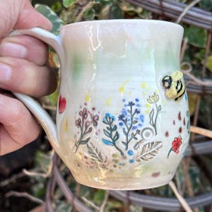 May include: A handmade ceramic mug with a white base, decorated with colourful floral designs and a 3D bee. The mug has a curved handle and is painted with a light green and yellow wash. A red heart is also painted on the mug.