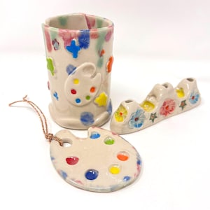 May include: A ceramic set featuring a cylindrical brush holder with a raised paint palette motif, a long brush rest with floral accents, and a hanging paint palette ornament with a copper string, all decorated with colourful paint splatters.