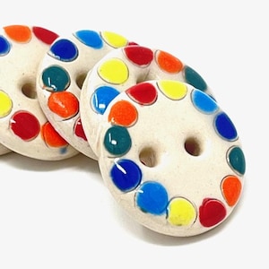 May include: Close-up of several off-white ceramic buttons with colourful, glossy dots in red, orange, yellow, green, and blue. Each button has two holes in the centre. The buttons are arranged in a slightly overlapping pattern.