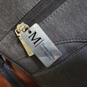 May include: Close-up of a silver rectangular tag with the letter "M" and the name "Thomas Marks" and phone number (234) 555-1212. The tag is attached to a black bag with a zipper.