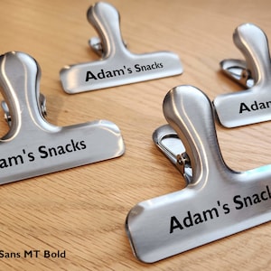 May include: Four silver metal clips with the text "Adam's Snacks" engraved in black. The clips are on a wooden surface.