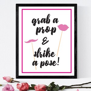 May include: A framed print with the text "grab a prop & strike a pose!" in black script. The print has a pink border and includes pink mustache and lips props. The frame is black. Roses are scattered at the bottom.