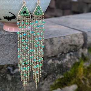 May include: Gold-tone earrings with a triangular top and cascading beaded strands. The triangle is filled with green beads, and the strands feature a mix of green and blue beads. The earrings are hanging in front of a stone wall.