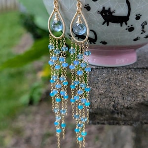 May include: Gold-toned dangle earrings with a teardrop design. Each earring features a large, light blue gemstone at the top, with cascading strands of small blue beads and gold chain. The earrings are displayed against a blurred green background.