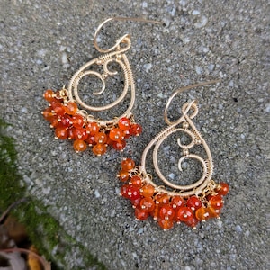 May include: A pair of gold-coloured wire earrings with a teardrop design. The earrings have a cluster of small, orange-red beads at the bottom. The wire is intricately looped and curled, creating an elegant and detailed appearance.