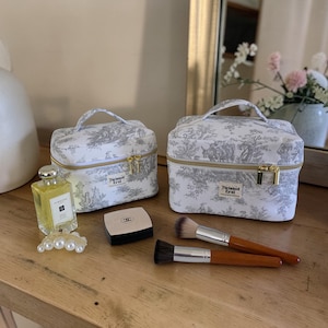 May include: Two white toiletry bags with a gray pastoral design and gold zippers sit on a wooden surface. A perfume bottle, a compact, and two makeup brushes are also on the surface. The bags have the text "Twisted Trail".