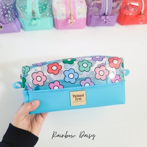 May include: A light blue pencil case with a clear top featuring a rainbow daisy print. The case has a brown label with the text "Twisted Erzi" on it. The text "Rainbow Daisy" is written in white below the case.