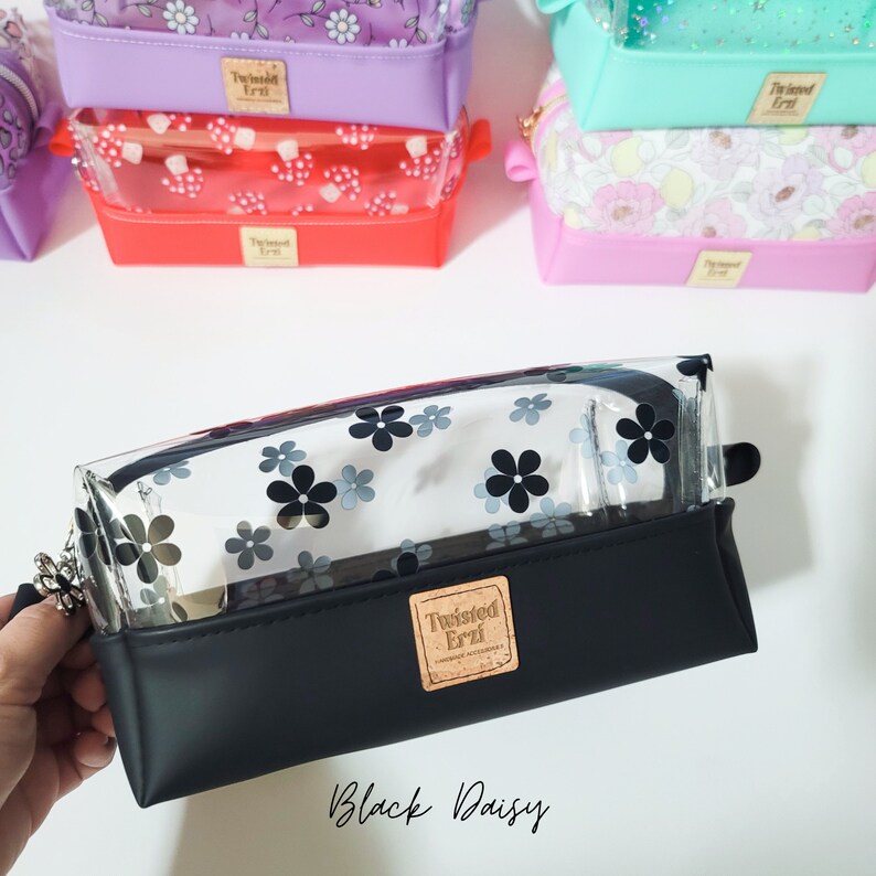 May include: A clear plastic pencil case with a black daisy print and a black leather base. The case has a zip closure and a Twisted Erzi logo.
