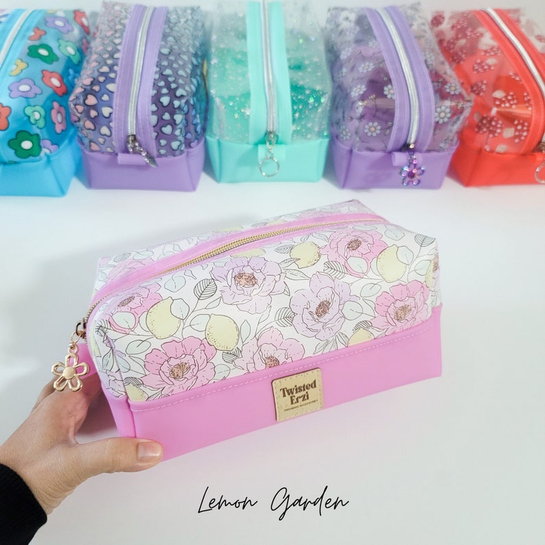 May include: A pink and white zippered pouch with a lemon and floral print. The pouch has a gold zipper and a gold charm with a flower design. The pouch has a pink bottom and a white top with the text "Twisted Erzi" printed on the bottom. The text "Lemon Garden" is printed below the pouch.