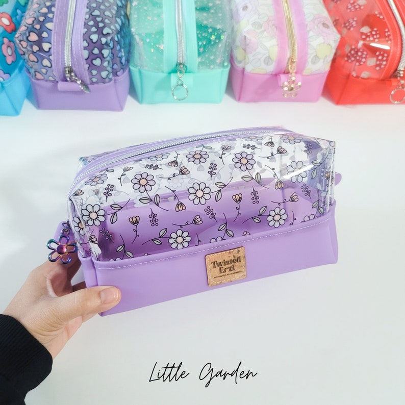 May include: A clear purple cosmetic bag with a floral print and a zipper closure. The bag has a clear plastic window and a purple fabric lining. The bag is labelled "Twisted Erzi" and "Little Garden".