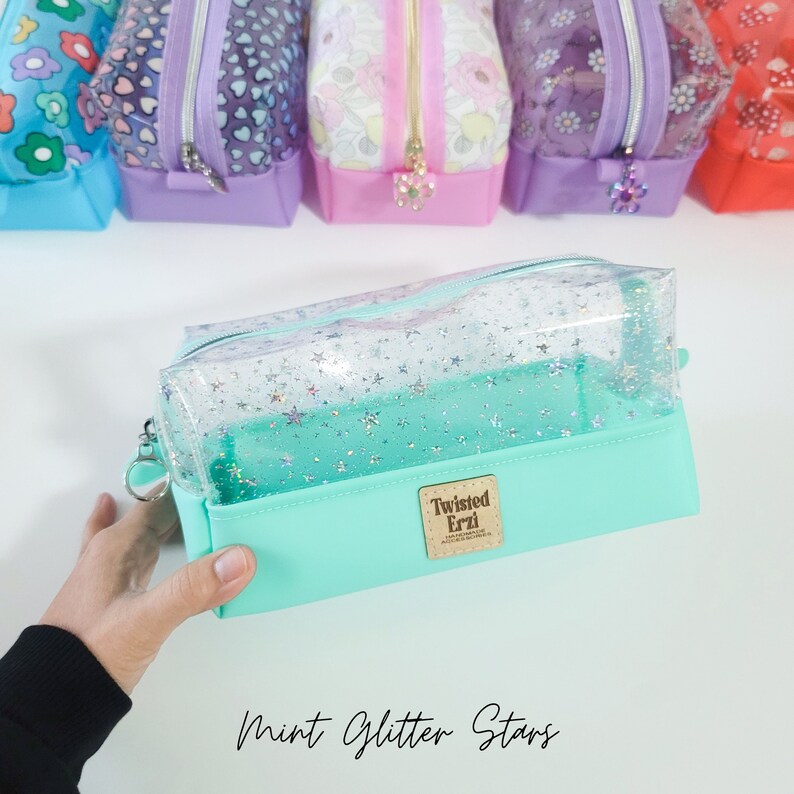 May include: A clear mint green pencil case with a glittery star pattern. The case has a zipper closure and a small brown leather tag with the text "Twisted Erzi" on it.