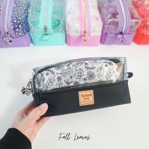 May include: A clear makeup bag with a black base and a white and black print of autumn leaves. The bag has a zip closure and a gold metal charm. The bag is labelled "Twisted Erzi" and "Fall Leaves".
