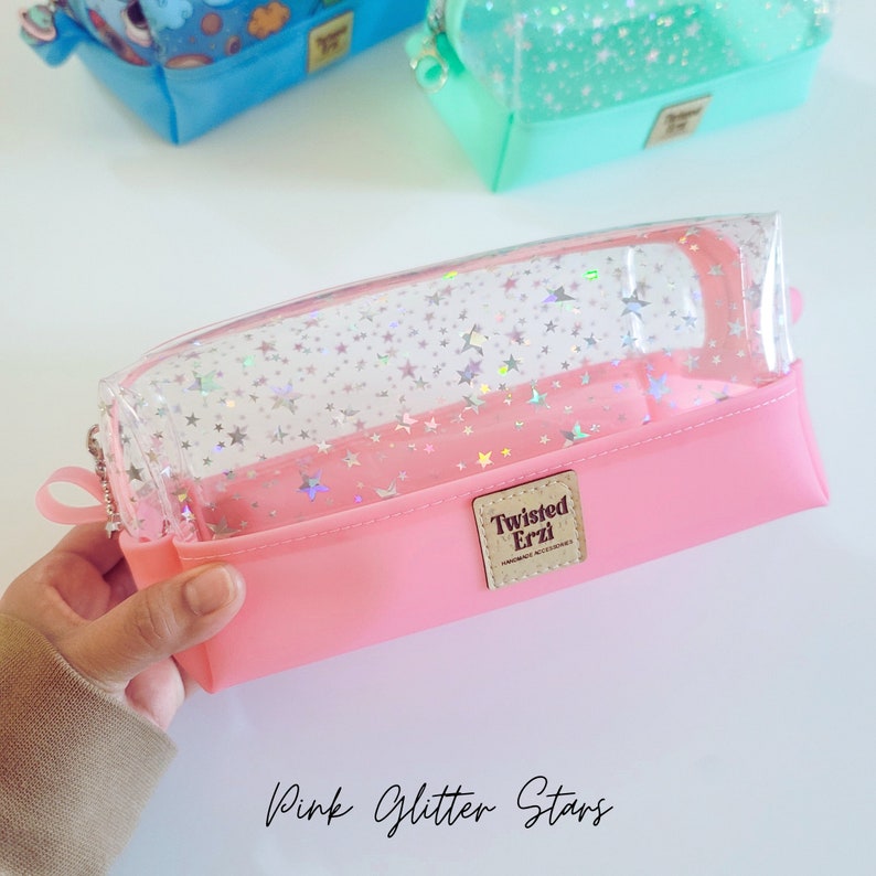 May include: Pink clear pencil case with a zipper closure and a clear top. The case is decorated with silver glitter stars. The case has a brown label with the text "Twisted Erzi" on it.