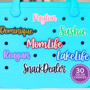 May include: A turquoise tote bag with various name and phrase decals in different colors. The decals include names like "Payton," "Dominique," and phrases like "Momlife" and "Snack Dealer." A circular sticker indicates "30 COLOR CHOICES."