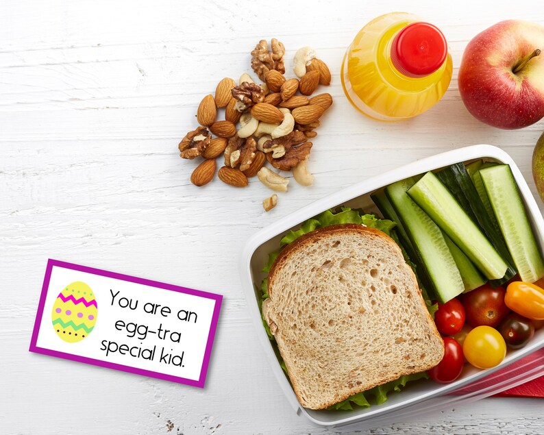 May include: A purple and white card with a yellow egg and the text "You are an egg-tra special kid."  A lunchbox with a sandwich, cucumber, tomatoes, and lettuce.  A red apple, a glass of orange juice, and a bowl of nuts are on a white wooden surface.