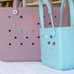 K&ouml;nnte beinhalten: Zwei Tragetaschen, eine hellviolette und eine hellblaue, mit passenden Griffen. Die Taschen haben eine Reihe runder L&ouml;cher und farbenfrohe dekorative Anstecknadeln. Die hellblaue Tasche befindet sich vor der hellvioletten Tasche.