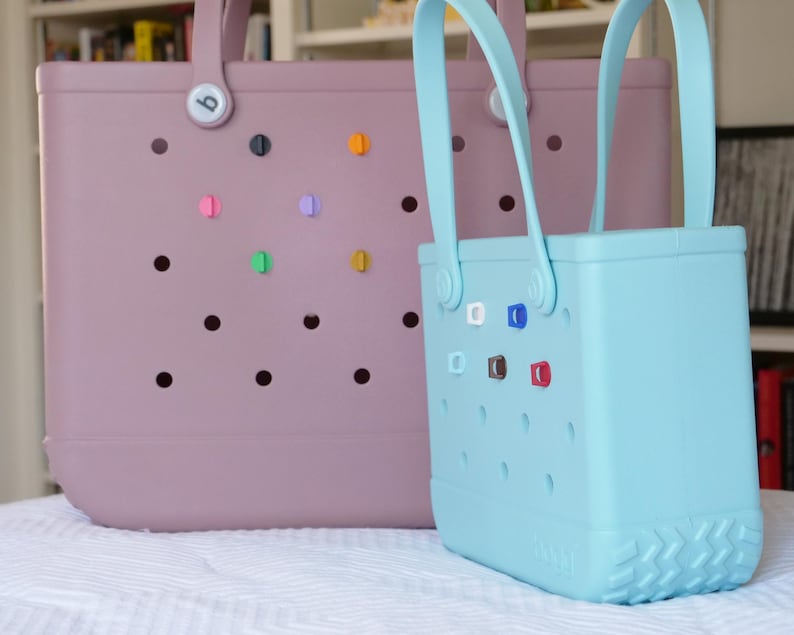 K&ouml;nnte beinhalten: Zwei Tragetaschen, eine hellviolette und eine hellblaue, mit passenden Griffen. Die Taschen haben eine Reihe runder L&ouml;cher und farbenfrohe dekorative Anstecknadeln. Die hellblaue Tasche befindet sich vor der hellvioletten Tasche.