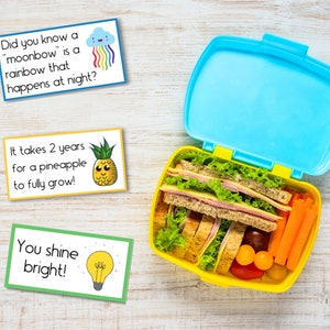 Puede incluir: Una fiambrera amarilla con tapa azul, llena de un s&aacute;ndwich, zanahorias y tomates cherry. Tres tarjetas coloridas con mensajes de &aacute;nimo y datos curiosos est&aacute;n sobre una superficie de madera.