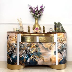 May include: A painted cabinet with a landscape design featuring trees, mountains, and water. The cabinet has two drawers with gold leaf accents and a curved shape. A gold vase with purple flowers and other decorative items sit on top. The cabinet is approximately 120 cm wide.