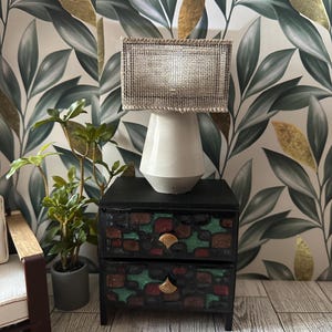 May include: A miniature lamp with a rectangular woven lampshade sits atop a white and gray ceramic base. The lamp rests on a small black nightstand with two drawers, decorated with red, green, and gold accents. A potted plant and a patterned wallpaper are in the background.