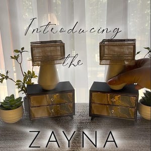 May include: Two small black chests with two drawers each, featuring a gold and green marbled front panel and silver knobs. Each chest supports a white vase topped with a rectangular gold mesh lampshade. Text reads: Introducing the ZAYNA COLLECTION.