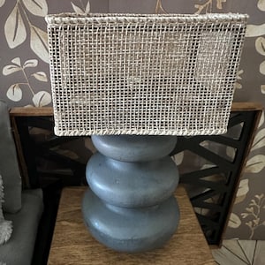 May include: A decorative table lamp with a gray stone base and a rectangular woven shade. The lamp sits on a wooden nightstand. The background features a floral wallpaper.