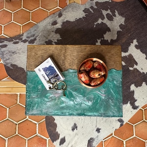 May include: A decorative wooden tray with a turquoise resin wave design. The tray holds a small copper bowl with decorative beads, a small book with the word "JOURNEY" on it, and a set of keys.
