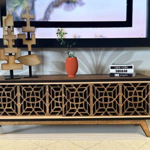 May include: A miniature wooden media console featuring two sliding doors with a geometric lattice pattern. The unit has a dark brown finish, four angled legs, and sits beneath two abstract wooden sculptures, a small orange vase, and three stacked books labeled New York, Chicago, and Los Angeles.