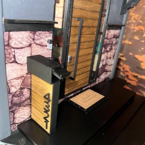 May include: A miniature model of a townhouse entrance. The wooden door has a long, silver handle and is framed by black trim. A welcome mat is visible in front of the door. The model features a stone wall and a black mailbox with the word "AKUA" on it.