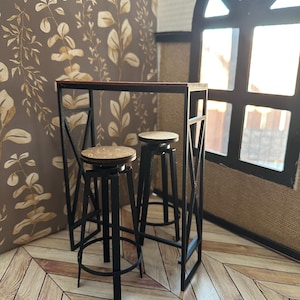 May include: A miniature black metal bar table with two matching stools. The table has a dark wood top and the stools have round, light-colored seats. The set is placed in a room with a brown and beige leaf-patterned wallpaper and a wooden floor.