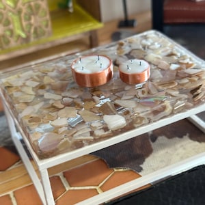 May include: A small, white-framed table with a clear resin top embedded with shell fragments. Two orange candles with multiple wicks sit on the table. The table rests on a patterned surface.