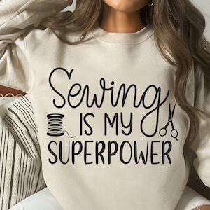 Puede incluir: Una sudadera beige con el texto "Coser es mi superpoder" en letras negras. El texto está acompañado por una ilustración negra de un carrete de hilo y un par de tijeras.