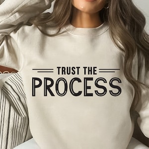 May include: A cream-colored sweatshirt with the text "TRUST THE PROCESS" printed in black.
